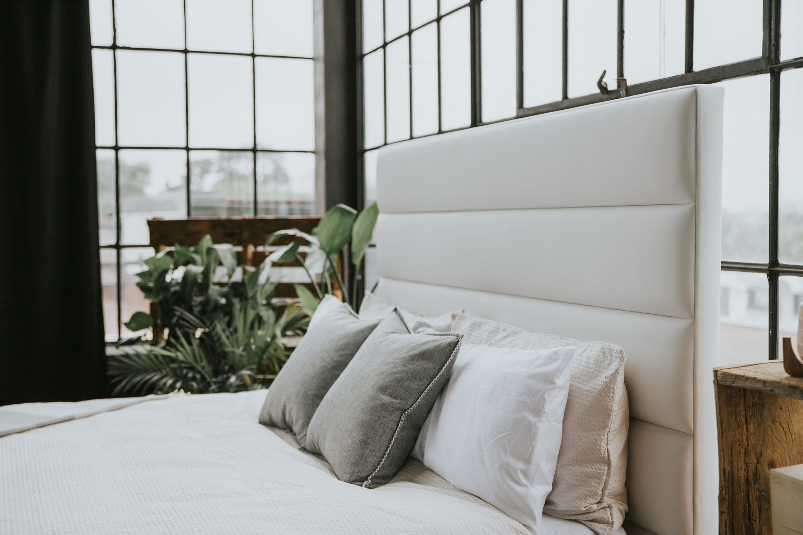 Layered grey and white pillows and a textured waffle-weave coverlet on a white upholstered bed in front of tall loft windows