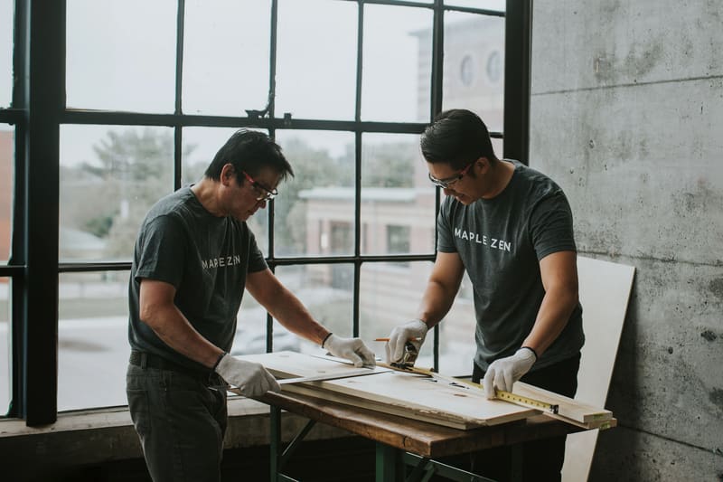 Two craftsmen in Maple Zen shirts measuring boards on a workbench in front of a tall window