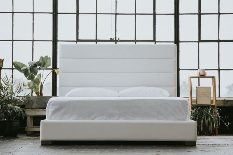 A white vegan leather platform bed with a horizontal channel headboard, straight on, in a loft with tall windows