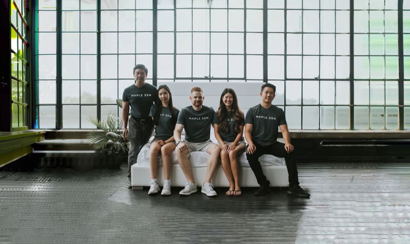 Five people in Maple Zen shirts around a white upholstered bed in front of tall loft windows